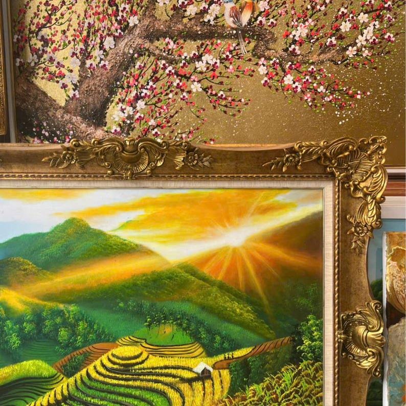 May include: A framed oil painting depicting a vibrant sunrise over green terraced rice fields and mountains, set within an ornate gold frame. Above it, a partial view of another painting shows a bird perched on a flowering tree branch.