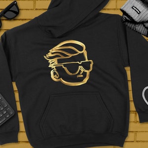 Puede incluir: Sudadera negra con un contorno dorado de un personaje de dibujos animados con gafas de sol. El personaje tiene un peinado grande y exagerado.