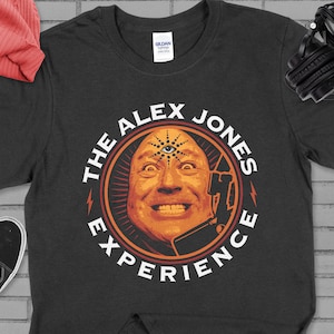 May include: Dark gray t-shirt featuring a graphic of a man's face with a third eye and the text "THE ALEX JONES EXPERIENCE" in a circular design. A red beanie and black sneakers are visible.