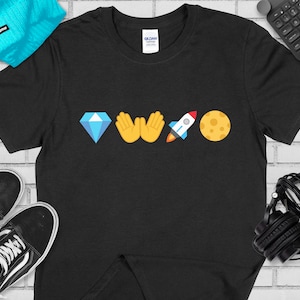 Könnte beinhalten: Schwarzes T-Shirt mit einem Diamanten, zwei erhobenen Händen, einer Rakete und einem Mond mit einem Gesicht darauf gedruckt.