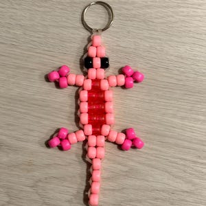 May include: A lizard-shaped keyring crafted from pink and red plastic pony beads. The lizard features a long tail, four legs with pink bead accents, and two solid black beads for eyes. A silver-toned metal split ring is attached to the top.