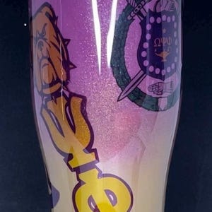 Può includere: Un bicchiere con glitter dorati e viola con un design a lettere greche e uno stemma di una confraternita. Lo stemma presenta uno scudo con una stella, una lampada e le lettere "ΩΦΔ".