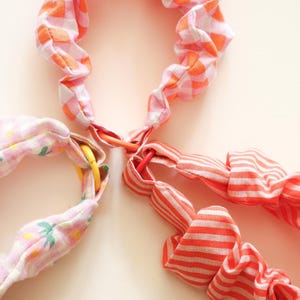 Peut inclure: Gros plan sur quatre bandeaux pour cheveux en tissu. Les bandeaux présentent différents motifs : vichy rose et orange, floral rose et rayures orange et blanches. Chaque bandeau est relié par un petit anneau coloré.