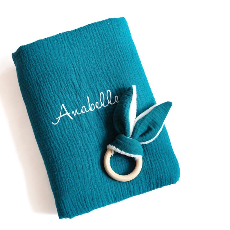 May include: A teal blue muslin swaddle blanket with the name "Anabelle" embroidered on it. A white and teal blue bunny rattle with a wooden ring is on top of the blanket.