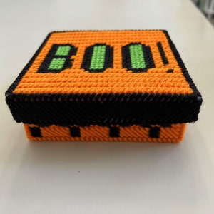 May include: Orange and black stitched box with the word "BOO!" in green and black. The box is square-shaped and appears to be handmade, perfect for Halloween decorations or storage.