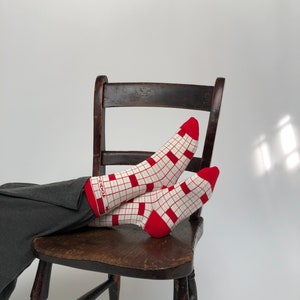 May include: White socks with a red grid design and red toes and cuffs. The socks are worn with dark grey trousers and are resting on a wooden chair. The socks have the text "100%" on the cuff.