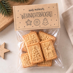 Könnte beinhalten: Ein durchsichtiger Plastikbeutel gefüllt mit rechteckigen Keksen, die mit Motiven von Schneeflocken, einem Weihnachtsbaum und einem Rentier geprägt sind. Der Beutel ist mit einem beigen Papieretikett verschlossen, das den Text aus meiner WEIHNACHTSBÄCKEREI trägt.