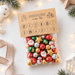 Puede incluir: Una bolsa de plástico transparente llena de chocolates navideños variados, incluyendo esferas envueltas en papel de aluminio con diseños de Santa Claus, muñecos de nieve, estrellas y copos de nieve. El encabezado de cartón dice ADVENTSKALENDER in der Tüte.