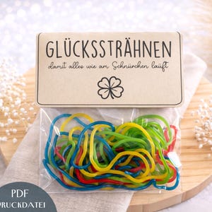 Könnte beinhalten: Ein klarer Plastikbeutel gefüllt mit bunten Gummibändern, darunter blau, gelb, grün und rot. Der Beutel ist versiegelt und auf einer Holzoberfläche platziert, mit einer dekorativen Karte oben. Die Karte hat Text und ein vierblättriges Kleeblatt.