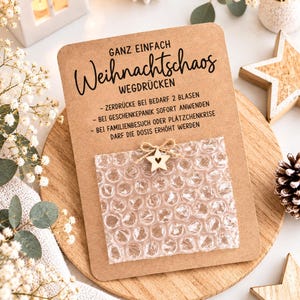 Könnte beinhalten: Eine rechteckige Karte aus Kraftpapier mit dem deutschen Text GANZ EINFACH Weihnachtschaos WEGDRÜCKEN, Anweisungen und einem quadratischen Stück Luftpolsterfolie in der Mitte, verziert mit einem kleinen Holzstern, der mit einer Schnur befestigt ist.