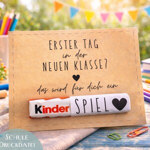 May include: A brown card with the text "ERSTER TAG in der NEUEN KLASSE? das wird für dich ein SPIEL" and a Kinder chocolate bar attached. The card is decorated with a black heart and the word "Kinder" is visible. The bottom of the card says "SCHULE DRUCKDATEI".