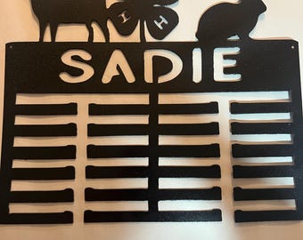 Puede incluir: Exhibidor de premios 4-H de metal negro con una silueta de cabra y conejo, un trébol 4-H y el nombre "SADIE" en la parte superior. El exhibidor tiene 16 ranuras para cintas.