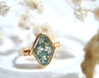 May include: A gold ring with a diamond-shaped green resin setting containing small white flowers and gold flakes.
