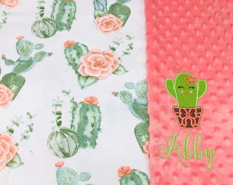 May include: A pink and white baby blanket with a cactus pattern on one side and a pink minky fabric on the other side. The minky side has an embroidered cactus with a smiling face and the name "Abby".