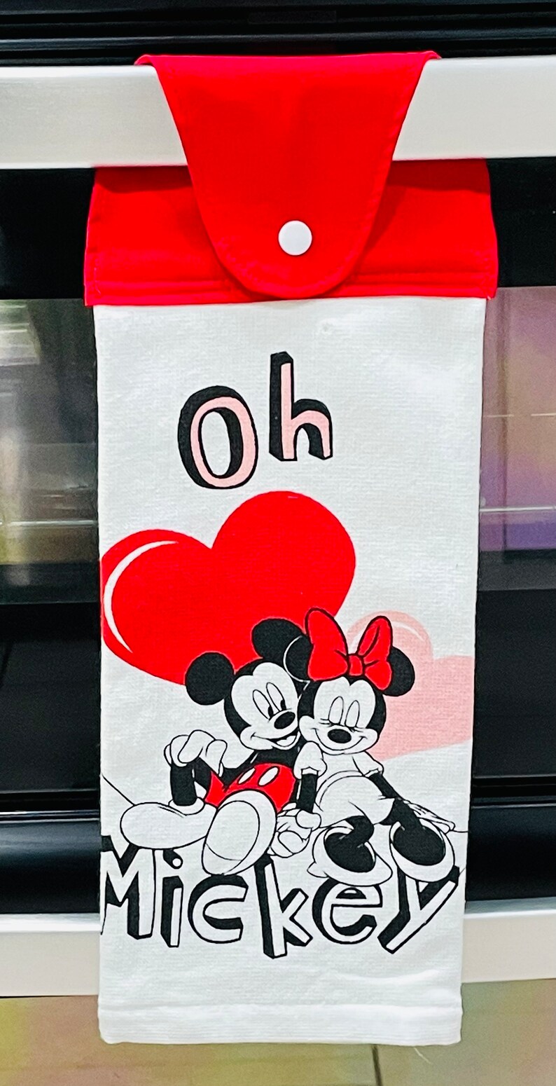 May include: White kitchen towel with a red heart, Mickey Mouse and Minnie Mouse, and the text "Oh Mickey". The towel has a red hanging loop with a white button.