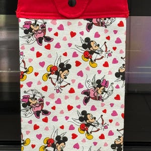 Puede incluir: Un pa&ntilde;o de cocina rojo y blanco con tem&aacute;tica de San Valent&iacute;n. El pa&ntilde;o presenta un patr&oacute;n de Mickey y Minnie Mouse vestidos de cupidos, junto con corazones rojos y rosas. La parte superior del pa&ntilde;o es roja con un bot&oacute;n negro.