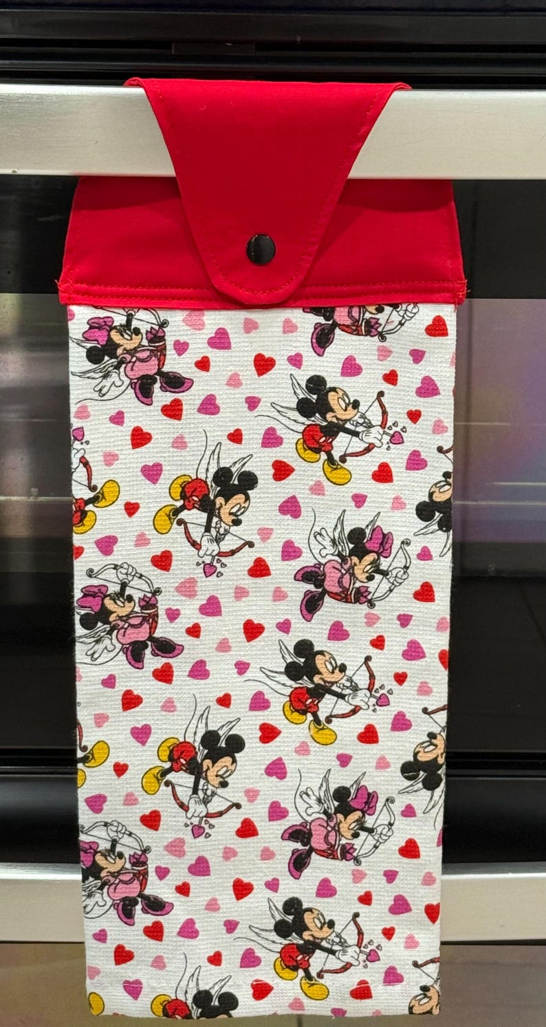 Puede incluir: Un pa&ntilde;o de cocina rojo y blanco con tem&aacute;tica de San Valent&iacute;n. El pa&ntilde;o presenta un patr&oacute;n de Mickey y Minnie Mouse vestidos de cupidos, junto con corazones rojos y rosas. La parte superior del pa&ntilde;o es roja con un bot&oacute;n negro.