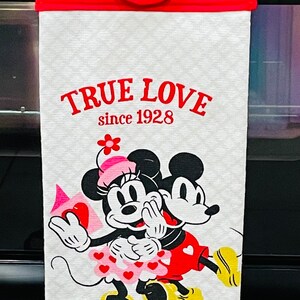 May include: White kitchen towel with a red hanging loop and a white button. The towel features a cartoon image of Mickey Mouse and Minnie Mouse with the text "TRUE LOVE since 1928".