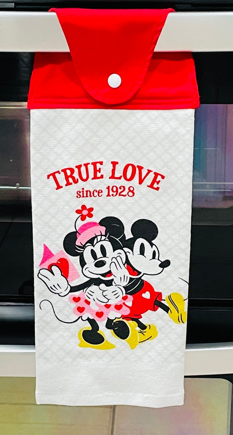 May include: White kitchen towel with a red hanging loop and a white button. The towel features a cartoon image of Mickey Mouse and Minnie Mouse with the text "TRUE LOVE since 1928".