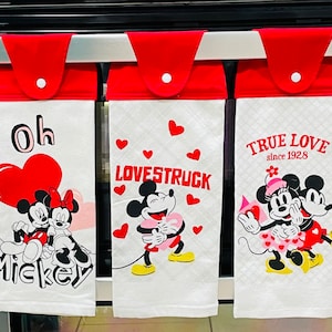 May include: Three white kitchen towels with red borders and Mickey Mouse themed designs. The first towel says "Oh Mickey" with a red heart and Mickey and Minnie Mouse. The second towel says "Lovestruck" with Mickey and Minnie Mouse. The third towel says "True Love since 1928" with Mickey and Minnie Mouse.
