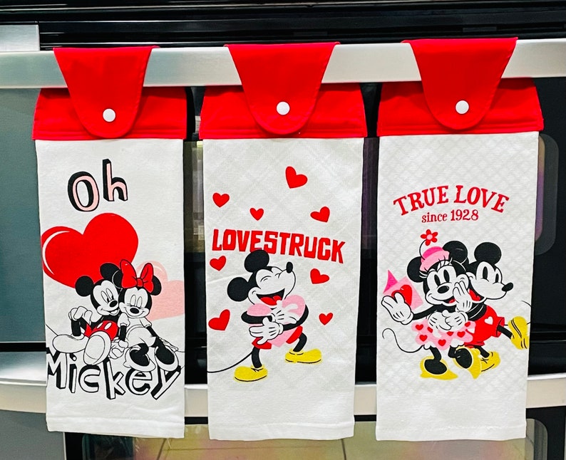May include: Three white kitchen towels with red borders and Mickey Mouse themed designs. The first towel says "Oh Mickey" with a red heart and Mickey and Minnie Mouse. The second towel says "Lovestruck" with Mickey and Minnie Mouse. The third towel says "True Love since 1928" with Mickey and Minnie Mouse.