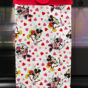 Puede incluir: Un pa&ntilde;o de cocina blanco con la parte superior roja, con un estampado de Mickey y Minnie Mouse como cupidos con arcos y flechas, rodeados de corazones rojos y rosas. El pa&ntilde;o est&aacute; colgado de una barra plateada.