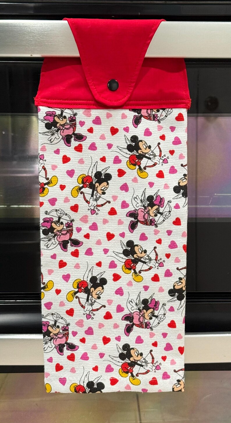 Puede incluir: Un pa&ntilde;o de cocina blanco con la parte superior roja, con un estampado de Mickey y Minnie Mouse como cupidos con arcos y flechas, rodeados de corazones rojos y rosas. El pa&ntilde;o est&aacute; colgado de una barra plateada.