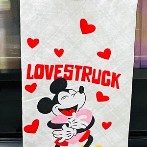 May include: White kitchen towel with a red hanging loop. The towel features a cartoon image of Mickey Mouse holding a pink heart and the text "LOVESTRUCK" in red letters. There are red hearts scattered around the image.