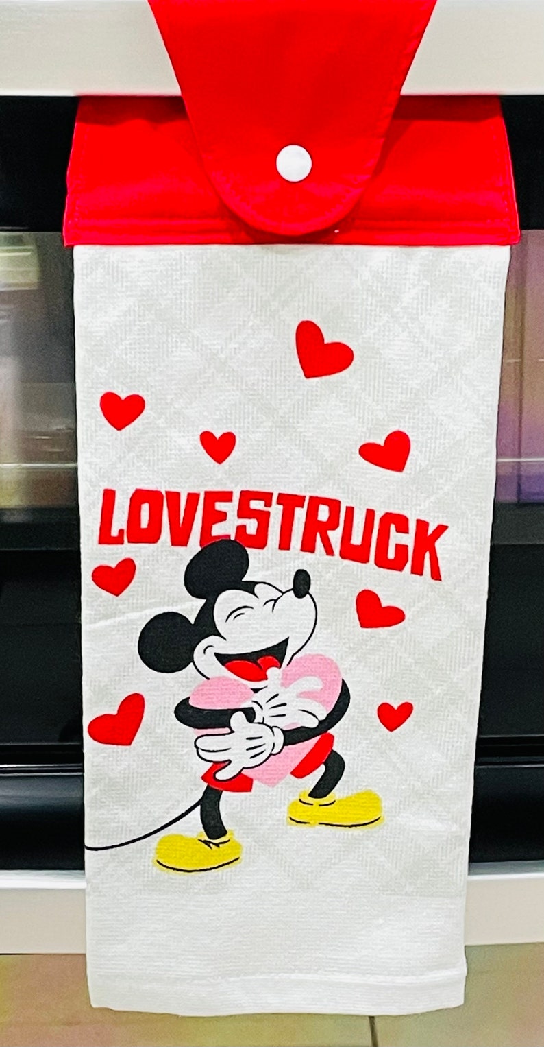 May include: White kitchen towel with a red hanging loop. The towel features a cartoon image of Mickey Mouse holding a pink heart and the text "LOVESTRUCK" in red letters. There are red hearts scattered around the image.