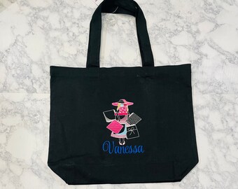 May include: Black canvas tote bag with an embroidered design of a woman in a hat holding shopping bags. The name "Vanessa" is embroidered below the design.