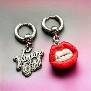May include: A pair of silver hoop earrings with charms. One charm is a red, sculpted, open mouth with white teeth. The other charm is a silver charm with the words "Vampire Girl" in a stylized font.