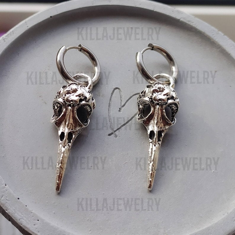 May include: A pair of silver hoop earrings with a bird skull charm. The skull has a long, pointed beak and is detailed with eye sockets and a textured surface.