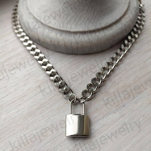 K&ouml;nnte beinhalten: Eine silberne Kettenkette mit einem silbernen Vorh&auml;ngeschloss-Anh&auml;nger. Die Kette ist im Curb-Stil.