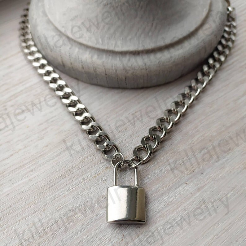 K&ouml;nnte beinhalten: Eine silberne Kettenkette mit einem silbernen Vorh&auml;ngeschloss-Anh&auml;nger. Die Kette ist im Curb-Stil.