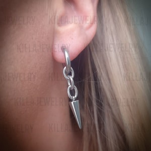 Może przedstawiać: Srebrny kolczyk z małym, spiczastym kolcem wiszącym na łańcuszku. Kolczyk jest noszony w górnym płatku ucha osoby.