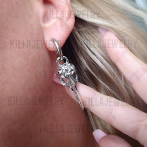 May include: Silver skull earring with a hoop and a dangling skull charm. The skull has a detailed design and a pointed beak.