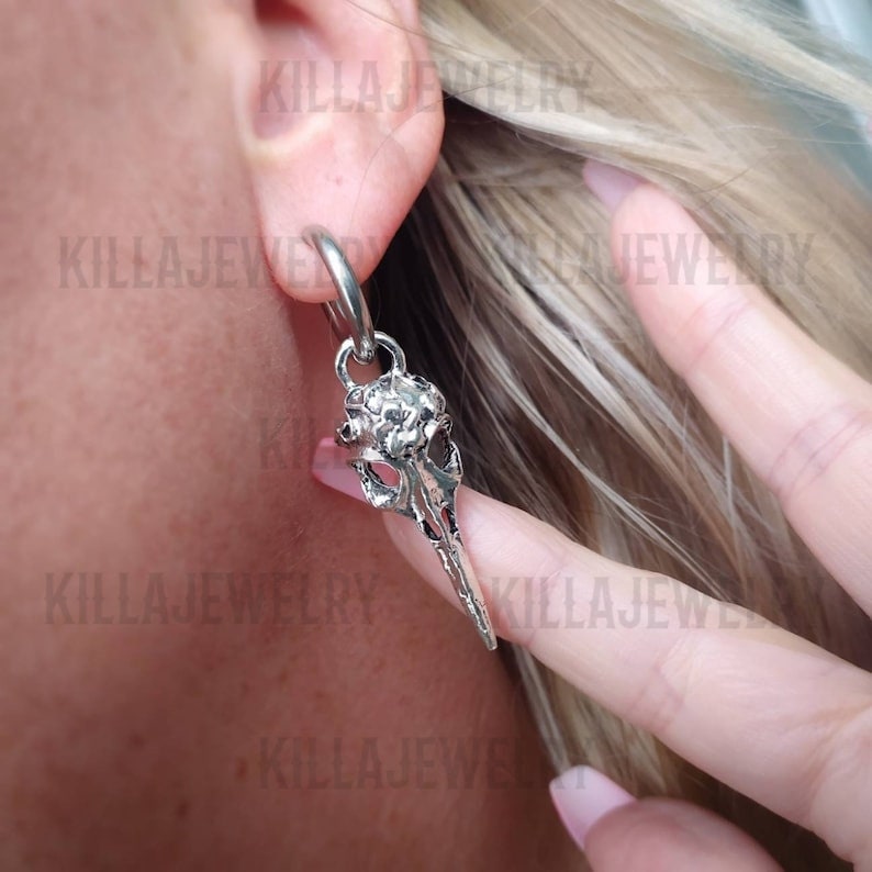 May include: Silver skull earring with a hoop and a dangling skull charm. The skull has a detailed design and a pointed beak.