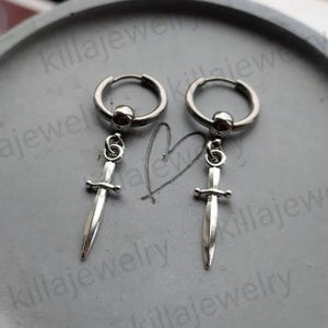 May include: A pair of silver hoop earrings with a silver sword charm dangling from each hoop.