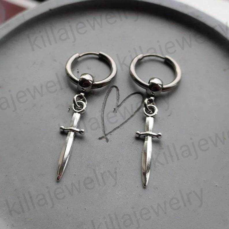 May include: A pair of silver hoop earrings with a silver sword charm dangling from each hoop.