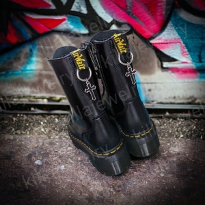 May include: Black leather platform boots with yellow stitching and a zipper. The boots have a silver cross charm and the words "AirWair" in yellow. The boots are on a concrete surface with graffiti in the background.