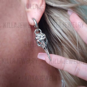 May include: Silver skull earring with a textured surface. The earring is attached to a hoop and hangs from the earlobe.