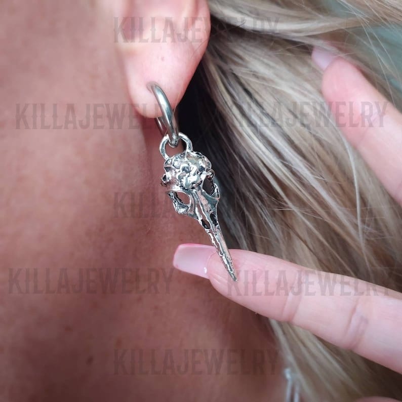 May include: Silver skull earring with a textured surface. The earring is attached to a hoop and hangs from the earlobe.