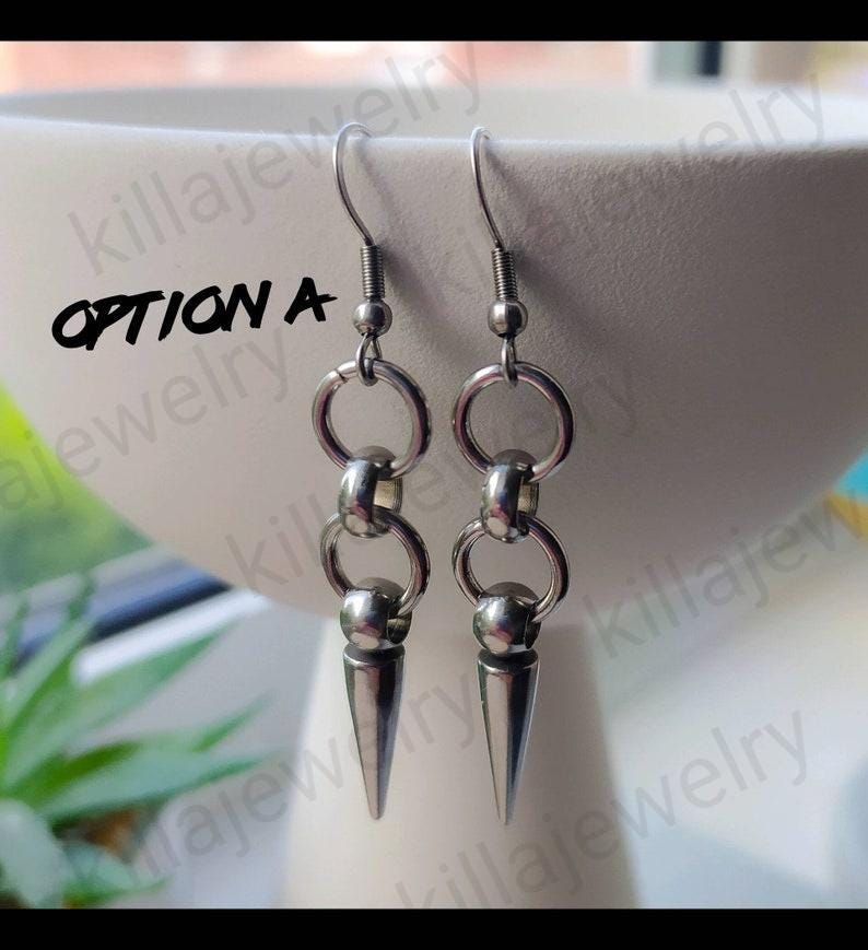 May include: A pair of silver earrings with a spike design. The earrings feature two connected rings and a small ball at the top of the spike.