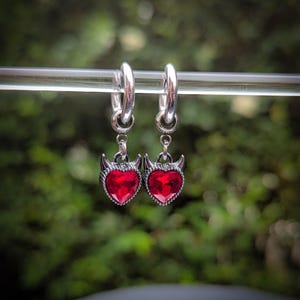 May include: Silver hoop earrings with red heart-shaped pendants. Each pendant has a devil horn design. The earrings are displayed against a blurred green backdrop. The earrings are a stylish accessory.