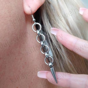 May include: Silver chain earrings with a pointed spike at the end. The earrings are made of metal and have a simple design.