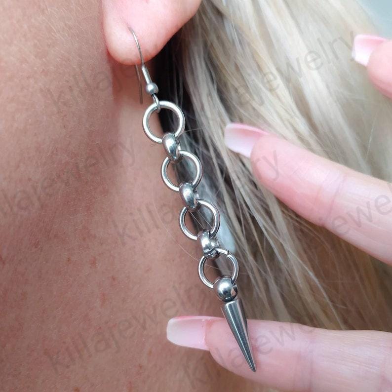 May include: Silver chain earrings with a pointed spike at the end. The earrings are made of metal and have a simple design.