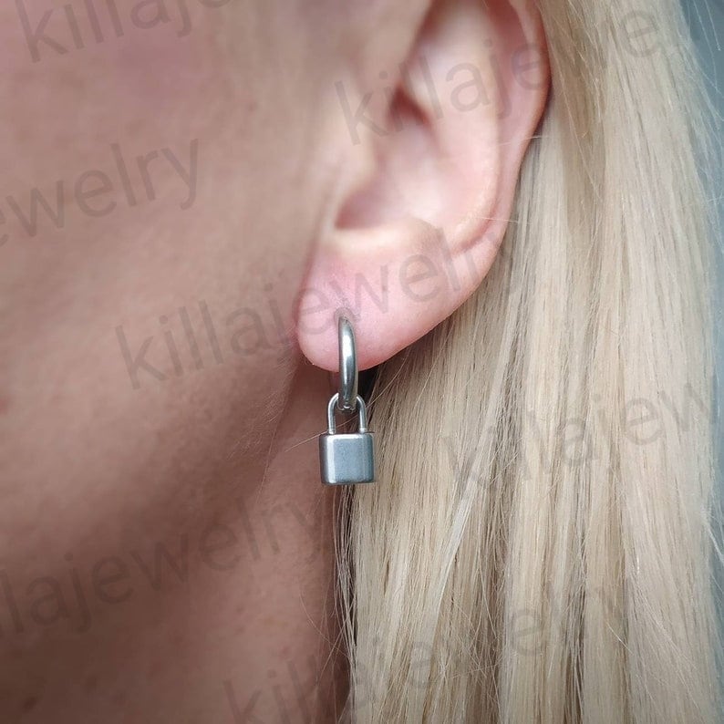 May include: A pair of silver padlock earrings with a hoop design. The earrings are worn in a person's ear.