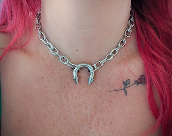 May include: A silver-colored chain necklace with a horseshoe-shaped pendant featuring two pointed spikes. The necklace is worn around the neck and is a bold, edgy accessory. The chain links are oval-shaped.