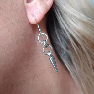 May include: Silver dangle earrings with a spike charm and two circular charms. The earrings are made of stainless steel.