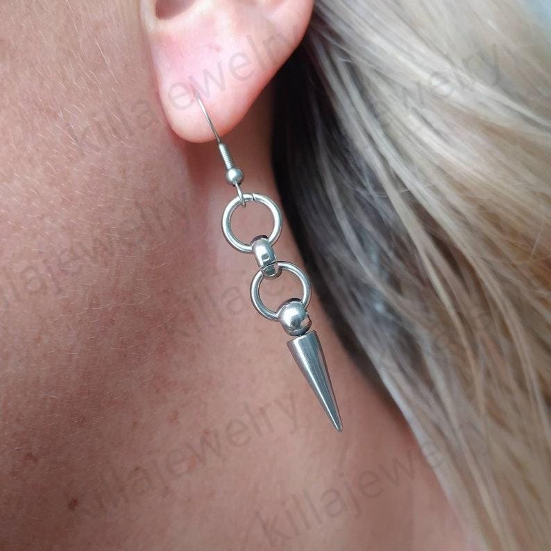 May include: Silver dangle earrings with a spike charm and two circular charms. The earrings are made of stainless steel.